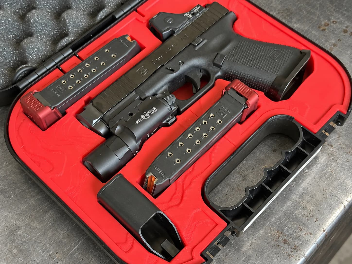 A 3D-printed red insert in a Glock factory case holding a Glock 19 with a red-dot optic and a Surefire light, two magazines with red basepads, and a spare part